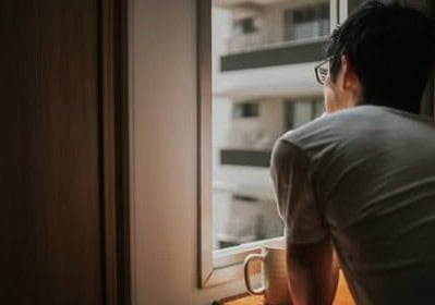 young man looking outside high rise apartment building window with cup of coffee