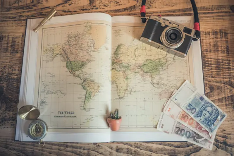World map with a camera, compass, and international currency on a wooden table